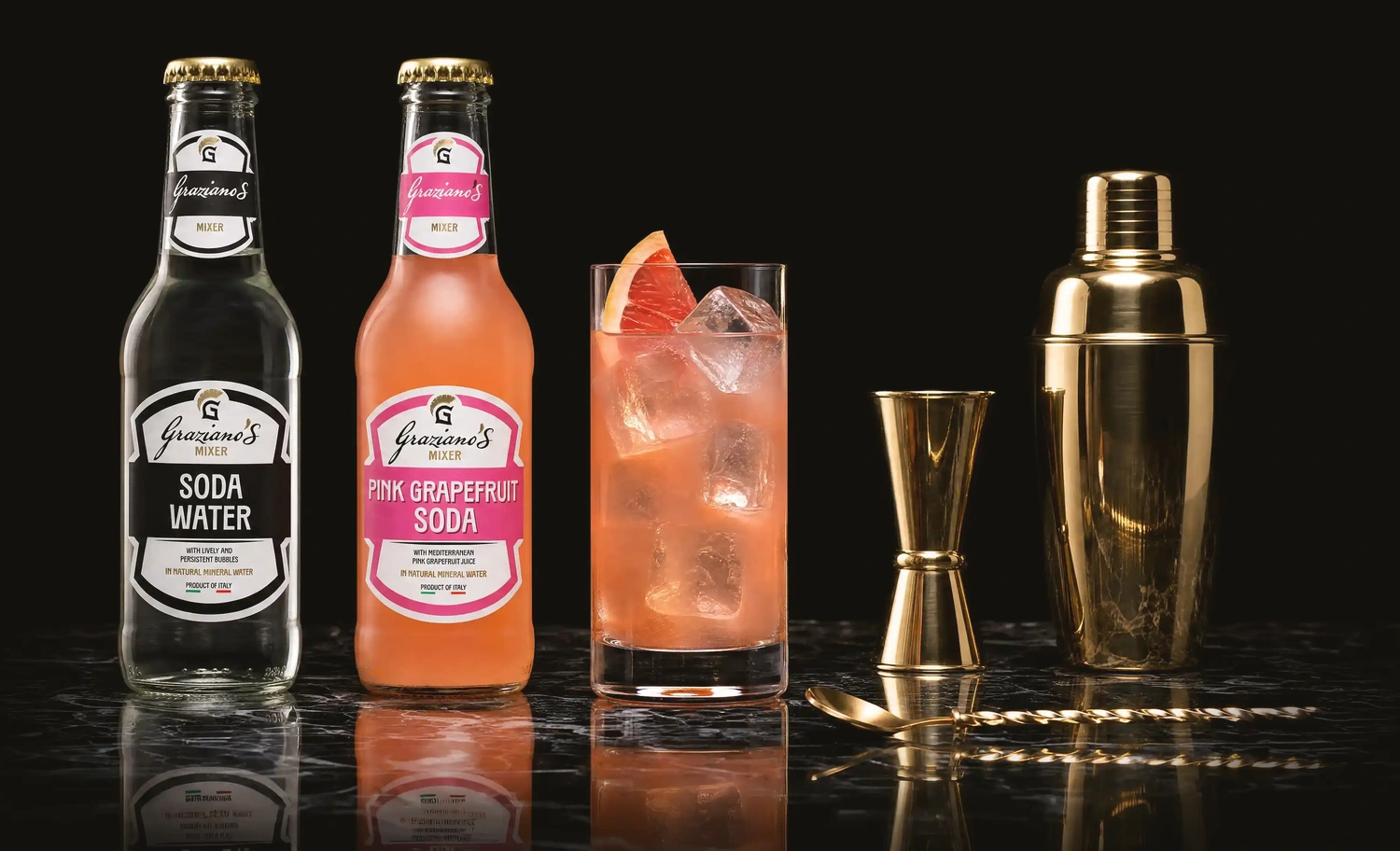 Graziano's Soda Water and Pink Grapefruit Soda bottles with a pink grapefruit cocktail, gold jigger and shaker on a cocktail bar counter